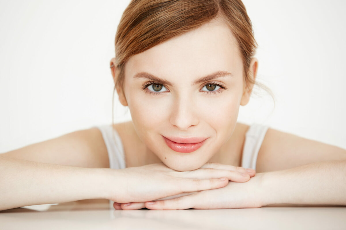 Beautiful girl with perfect clean skin smiling looking at camera sitting at table over white background. Beauty spa and cosmetology.
